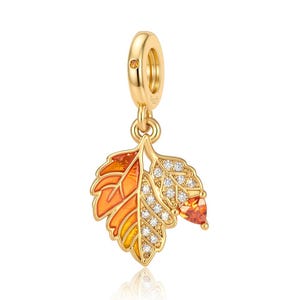 May include: A gold-toned charm featuring two leaves hanging from a circular bail. One leaf is filled with orange and yellow enamel, while the other is encrusted with clear cubic zirconia stones and a single triangular orange stone at the tip.