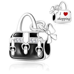 May include: A silver charm shaped like a handbag with black enamel detailing, clear stones, and a dangling bow. A smaller view shows the back of the charm with the text I heart shopping, featuring a red heart symbol, engraved on the silver surface.