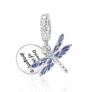 Op de afbeelding: Een zilveren libellenbedel met blauwe emaille vleugels en een kleine, ronde, lichtblauwe edelsteen in het midden. De bedel heeft een ronde hanger met de woorden "Dance of the dragon" in zwarte tekst. De bovenste lus is versierd met kleine bloemen.
