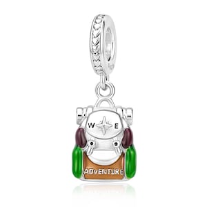 May include: A silver charm in the shape of a backpack featuring a compass rose design on the top flap, the word ADVENTURE printed on the front pocket, and green, brown, and dark red enamel accents. The bail is decorated with a repeating heart pattern.