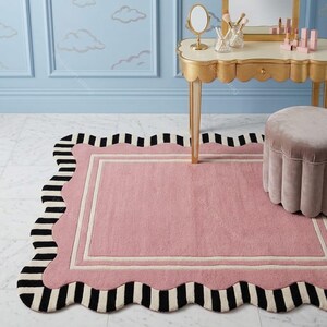 May include: A pink rectangular rug with a scalloped edge and a black and white striped border. The rug has a cream-colored inner border. The rug is placed in front of a vanity table and a small stool.