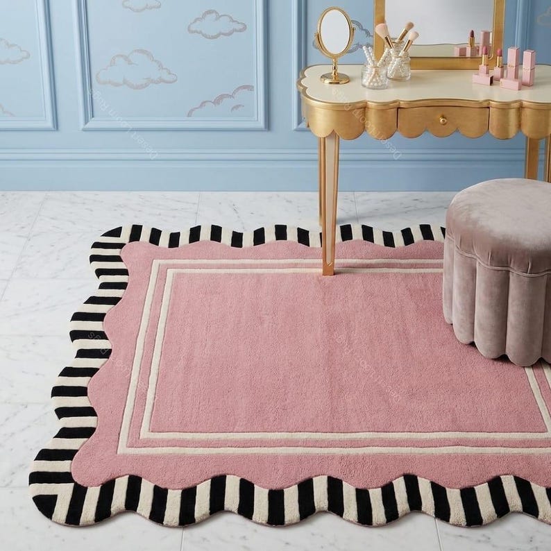 May include: A pink rectangular rug with a scalloped edge and a black and white striped border. The rug has a cream-colored inner border. The rug is placed in front of a vanity table and a small stool.