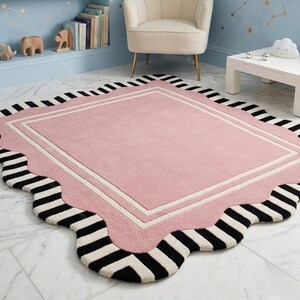 May include: A pink rug with a scalloped edge and a black and white striped border. The rug has a rectangular center with a white border. The rug is on a marble floor.