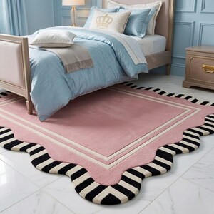 May include: A pink rug with a scalloped edge and a rectangular center. The rug features a black and white striped border and a white and pink inner border. The rug is placed in front of a bed with light blue bedding.