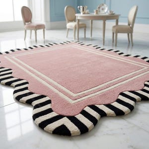 May include: A rectangular pink rug with a scalloped edge and a black and white striped border. The rug has a white and cream-colored inner border. The rug is in a room with light blue walls and white marble flooring.