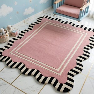 May include: A pink rectangular rug with a scalloped edge and a black and white striped border. The rug has a white border and is placed on a tiled floor. A blue and pink swing chair is in the background.