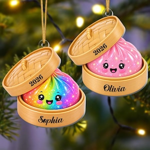 May include: Two hanging ornaments shaped like bamboo steamer baskets containing glittery, smiling dumpling characters. One dumpling is rainbow-colored with the name Sophia, the other is pink with the name Olivia. Both feature the year 2026 printed on the lid.