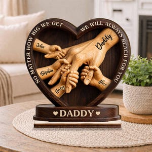 Könnte beinhalten: Eine herzförmige Holzskulptur mit dem Wort "Daddy" und den Namen John, Sophia, Henry und Alice. Die Skulptur zeigt Hände, die sich gegenseitig halten, mit dem Text "Egal wie groß wir werden, wir werden immer nach dir greifen."