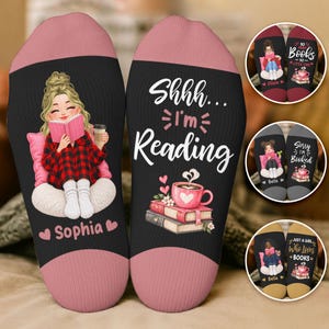 Pode incluir: Um par de meias pretas com punhos cor-de-rosa. Uma meia apresenta uma personagem loira a ler um livro com o nome Sophia por baixo. A outra exibe o texto Shhh... I'm Reading sobre uma pilha de livros e uma caneca rosa. Pequenos círculos mostram variações.