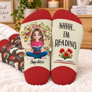 May include: A pair of cream-colored socks with red toe and heel sections. One sock features a cartoon girl reading under a tree with the name Sophia. The other sock displays the text SHHH... I'M READING above an open book with red poppies.