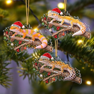 Könnte beinhalten: Drei flache Hängedekorationen mit Leopardengeckos auf Ästen, geschmückt mit Lichterketten, Tannenzapfen und roten Beeren. Jeder Gecko trägt eine rote Weihnachtsmütze mit den Namen Oliver, Johnny oder Lucian sowie der Jahreszahl 2026.