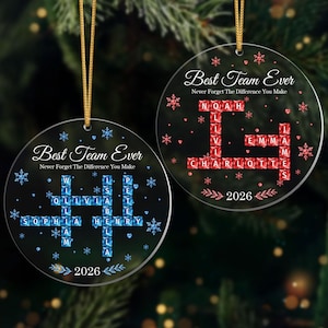 Op de afbeelding: Twee ronde, doorzichtige acryl ornamenten met gouden ophangkoorden. Eén toont blauwe kristalachtige lettertegels die namen vormen in een kruiswoordraadselpatroon met blauwe sneeuwvlokken en de tekst Best Team Ever Never Forget The Difference You Make 2026.