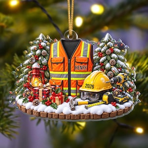 Op de afbeelding: Een decoratief ornament met een oranje veiligheidsvest met de naam HENRY, een gele veiligheidshelm met het opschrift 2026, een boormachine, een hamer, een rolmaat, een lantaarn en ingepakte cadeaus, tegen een achtergrond van besneeuwde dennen.