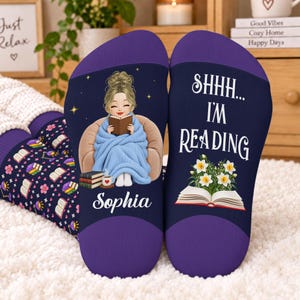 May include: A pair of dark blue socks with purple cuffs. One sock features a cartoon character reading a book while wrapped in a blue blanket, with the name Sophia printed below. The other sock displays the text SHHH... I'M READING above an open book with white flowers.