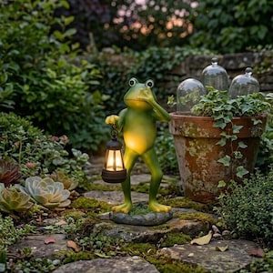 Puede incluir: Una estatua de rana verde sosteniendo una linterna negra con luz cálida. La rana está de pie sobre una piedra en un jardín, con plantas y una maceta de terracota en el fondo. La estatua tiene una expresión de sorpresa.