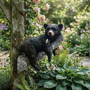 Peut inclure: Une statue d'ours noir se reposant sur une branche d'arbre, avec une petite abeille et un nid d'abeilles. L'ours a un nez rose et est entouré de verdure et de fleurs luxuriantes, créant un charmant élément de jardin.