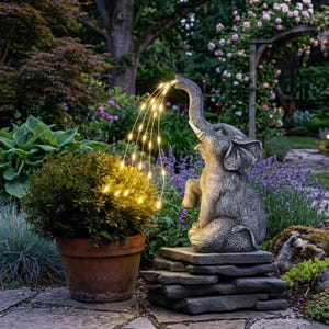 Könnte beinhalten: Eine graue Elefanten-Gartenstatue mit erhobenem Rüssel, die einen Strom warmweißer Lichterketten zu versprühen scheint. Die Statue steht auf einem Stapel Steinplatten, mit einer Topfpflanze links. Der Hintergrund zeigt eine üppige Gartenlandschaft.