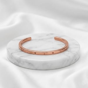 May include: A rose gold-toned cuff bracelet featuring a smooth, polished finish with evenly spaced vertical indentations and small circular depressions along the outer band, creating a structured and uniform appearance throughout the metal surface.