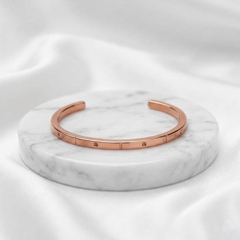 May include: A rose gold-toned cuff bracelet featuring a smooth, polished finish with evenly spaced vertical indentations and small circular depressions along the outer band, creating a structured and uniform appearance throughout the metal surface.