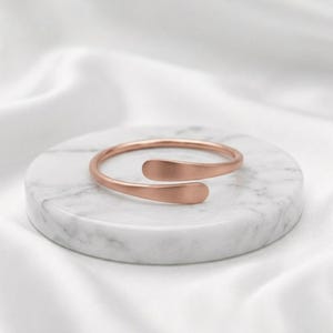 May include: A rose gold-toned open cuff ring with a smooth, matte finish. The ring features a bypass design where the two rounded ends overlap slightly, creating a continuous, minimalist circular band that is open at the front.