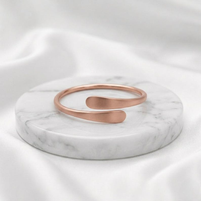 May include: A rose gold-toned open cuff ring with a smooth, matte finish. The ring features a bypass design where the two rounded ends overlap slightly, creating a continuous, minimalist circular band that is open at the front.