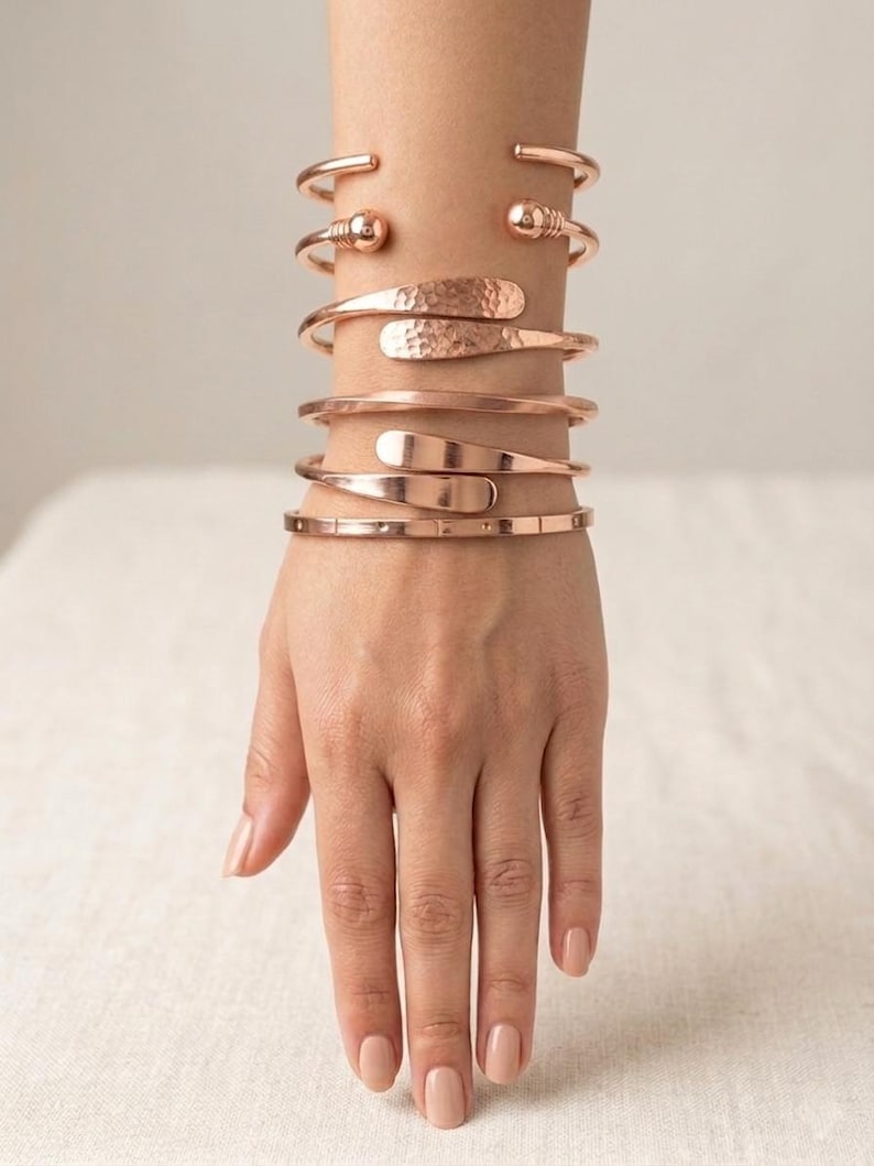May include: A set of four rose gold-toned metal cuff bracelets worn on the wrist. The top cuff features ball-end terminals, the second has a hammered texture with flat ends, the third is a smooth wrap, and the bottom is a thin, segmented band.