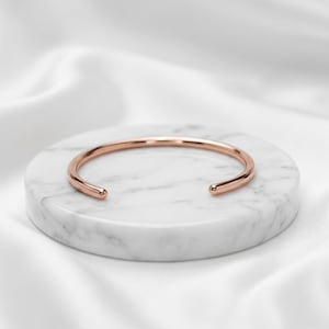 May include: A polished rose gold cuff bracelet with a smooth, rounded surface and an open design. The bracelet features a simple, continuous band that curves into a circular shape with two rounded ends, showcasing a metallic finish.