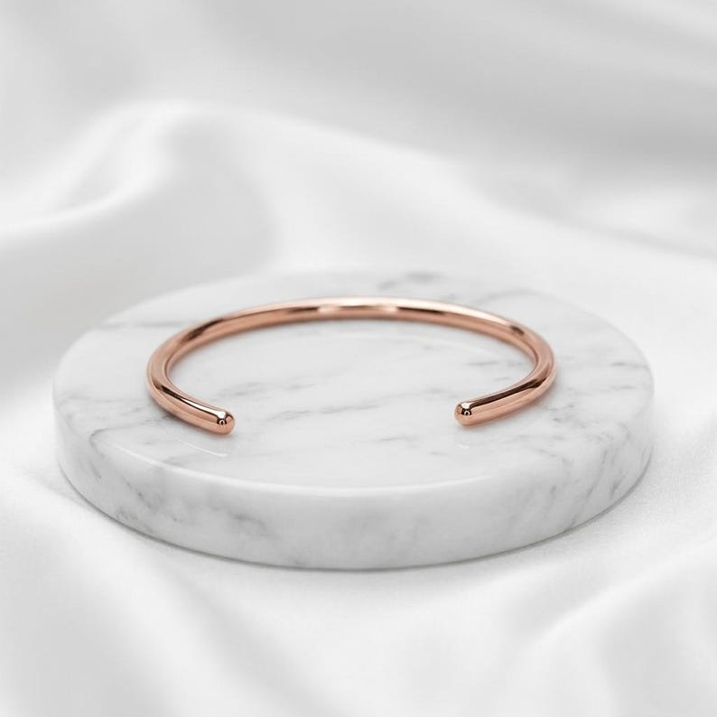 May include: A polished rose gold cuff bracelet with a smooth, rounded surface and an open design. The bracelet features a simple, continuous band that curves into a circular shape with two rounded ends, showcasing a metallic finish.