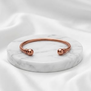 May include: A copper-toned metal cuff bracelet featuring a smooth, open-ended design. The bracelet ends in two rounded spheres, each accented with three thin, parallel engraved lines near the tips, creating a simple and polished appearance.