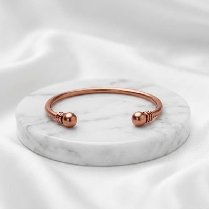 May include: A copper-toned metal cuff bracelet featuring a smooth, open-ended design. The bracelet ends in two rounded spheres, each accented with three thin, parallel engraved lines near the tips, creating a simple and polished appearance.