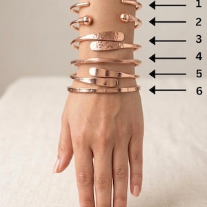 May include: Six copper cuff bracelets arranged in a vertical stack, numbered one through six with black arrows. The designs include plain bands, ball-end terminals, hammered textures, and smooth flat-end styles. The text Pure Coppe is visible.