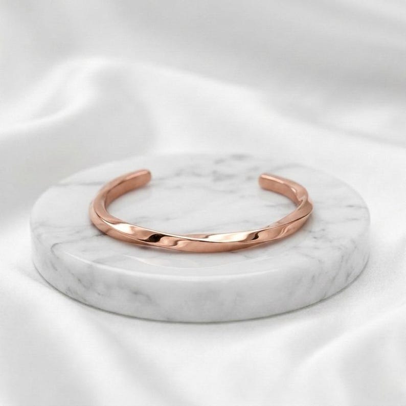 May include: A rose gold cuff bracelet with a smooth, twisted metal design. The bracelet features a polished, reflective surface and an open-ended circular shape, showcasing a continuous, flowing metallic form with a warm, pinkish-gold hue.