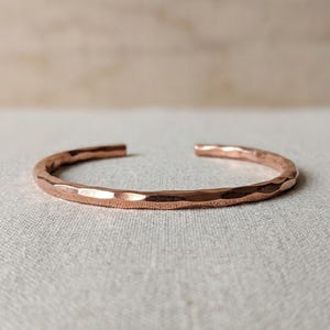 Op de afbeelding: Een koperen armband met een opvallende gehamerde textuur over het gehele buitenoppervlak. De metalen band is aan de uiteinden open en vormt een cirkelvorm met een gepolijste, reflecterende afwerking die het licht op de randen opvangt.