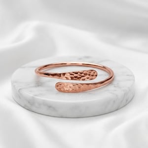May include: A rose gold cuff bracelet featuring a hammered texture across its entire surface. The bracelet has an open design with two rounded, slightly overlapping ends, creating a continuous circular shape with a polished metallic finish.