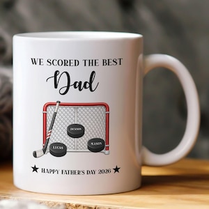 Op de afbeelding: Een witte keramische mok met een ijshockey-afbeelding met een rood doelnet, een hockeystick en drie zwarte pucks met de namen Jackson, Lucas en Mason. De tekst luidt WE SCORED THE BEST Dad en HAPPY FATHER'S DAY 2026 met twee zwarte sterren.