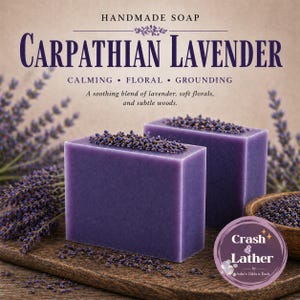 May include: Two square, purple handmade soaps with Carpathian Lavender text. The soaps are topped with dried lavender buds. The image also includes sprigs of lavender and a wooden bowl filled with lavender buds. The soap is labeled "Crash & Lather".