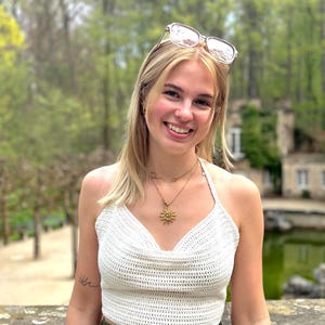 Puede incluir: Top de ganchillo blanco con diseño de cuello en V. El top se combina con pantalones verde oliva. También son visibles un collar con colgante de sol dorado y gafas de sol. El fondo presenta un parque con árboles y un edificio.