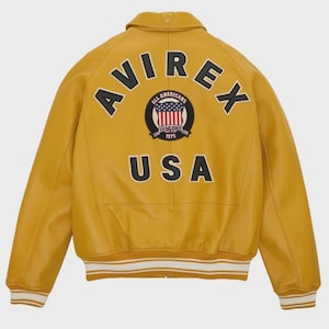 May include: Mustard yellow leather jacket with the word "AVIREX" arched across the back, above a circular patch with the words "ALL AMERICANS" and "USA". The jacket has white and cream striped cuffs and hem.