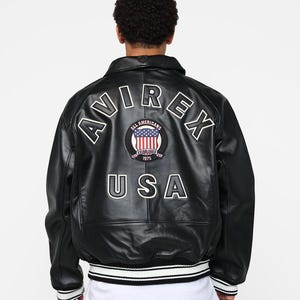 May include: Black leather bomber jacket with white lettering and a circular patch featuring the American flag. The jacket has a ribbed hem with white and black stripes and the text "AVIREX USA".