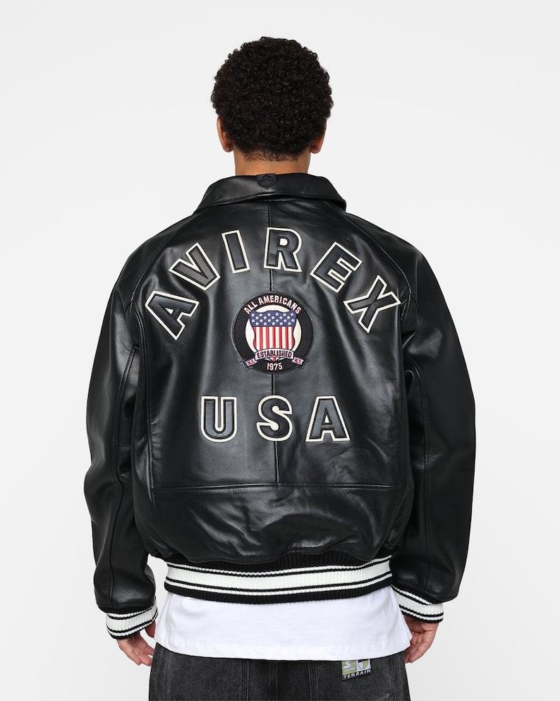 May include: Black leather bomber jacket with white lettering and a circular patch featuring the American flag. The jacket has a ribbed hem with white and black stripes and the text "AVIREX USA".