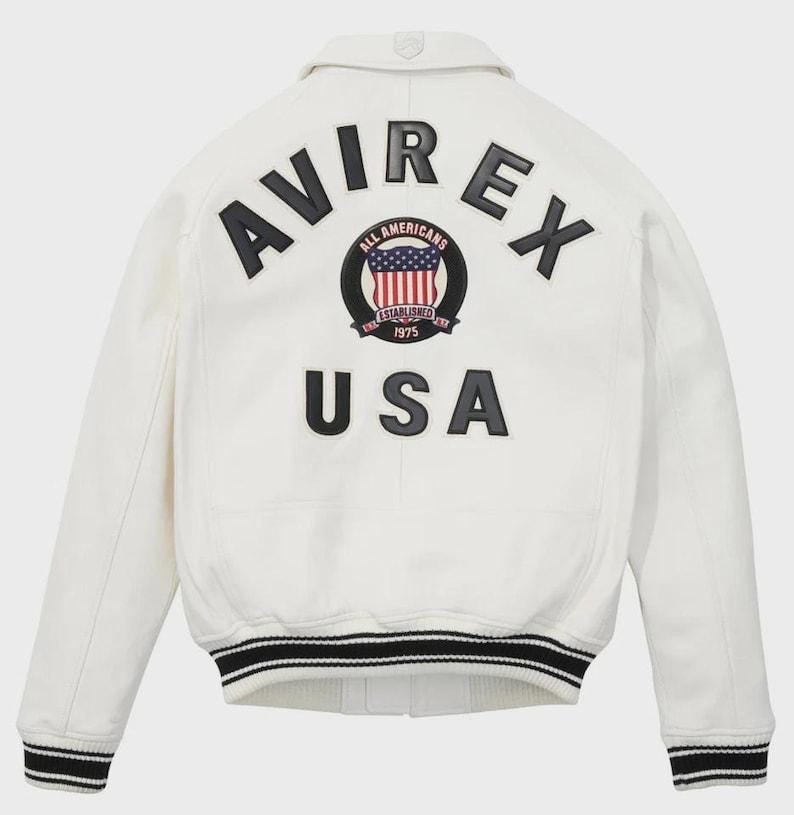 May include: White leather jacket with black and white striped trim at the cuffs and waist. The back features the text "AVIREX USA" in black lettering, with a circular emblem that includes the American flag and the words "ALL AMERICANS ESTABLISHED 1975".