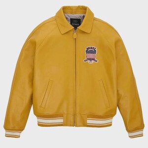 May include: Mustard yellow leather jacket with a collared neckline and a zipper closure. Features a patch with a patriotic design on the chest and white and gold striped cuffs and hem. The jacket has two front pockets and the brand name "AVIREX" is visible.