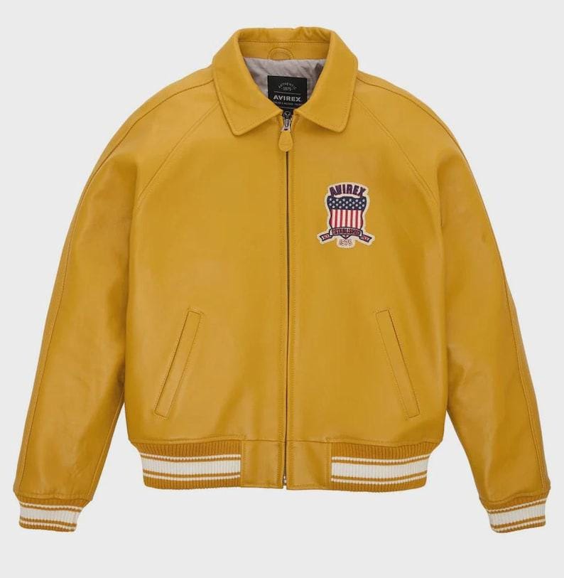 May include: Mustard yellow leather jacket with a collared neckline and a zipper closure. Features a patch with a patriotic design on the chest and white and gold striped cuffs and hem. The jacket has two front pockets and the brand name "AVIREX" is visible.