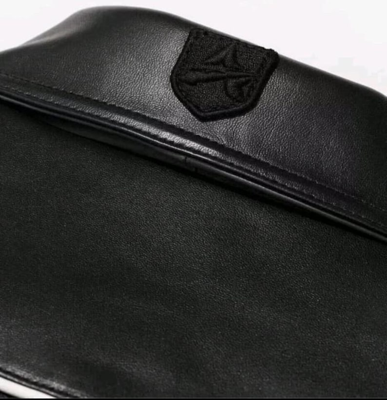 Op de afbeelding: Een zwart lederen kledingstuk met een gladde, matte textuur en een opvallende, gestikte zwarte patch op de kraag. De patch toont een verhoogd, in reli&euml;f gemaakt embleem met een geometrisch ontwerp, gecentreerd op de bovenrand van de kraag.