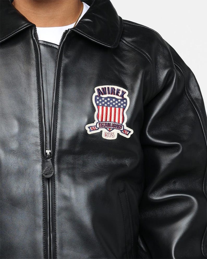 May include: Black leather jacket with a zip-up front and a collared neckline. Features a patch on the chest with the text "AIREX NY ESTABLISHED 1975" in red, white, and blue. The jacket has a classic, timeless design.