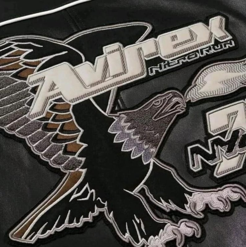 Op de afbeelding: Een zwart leren jack met een grote geborduurde adelaarspatch met bruine en grijze vleugeldetails. De tekst AVIREX is in het wit boven de adelaar gestikt, met NITRO RUN en NY eronder. Het ontwerp bevat witte omlijningen en dikke letters.