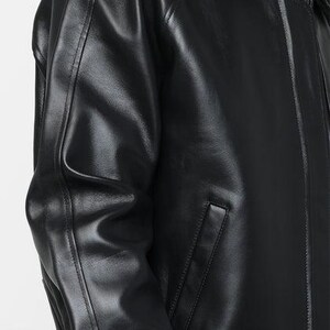 May include: A close-up of a black leather jacket. The jacket has a collar, a zipper, and a pocket on the front. The leather appears shiny and smooth.