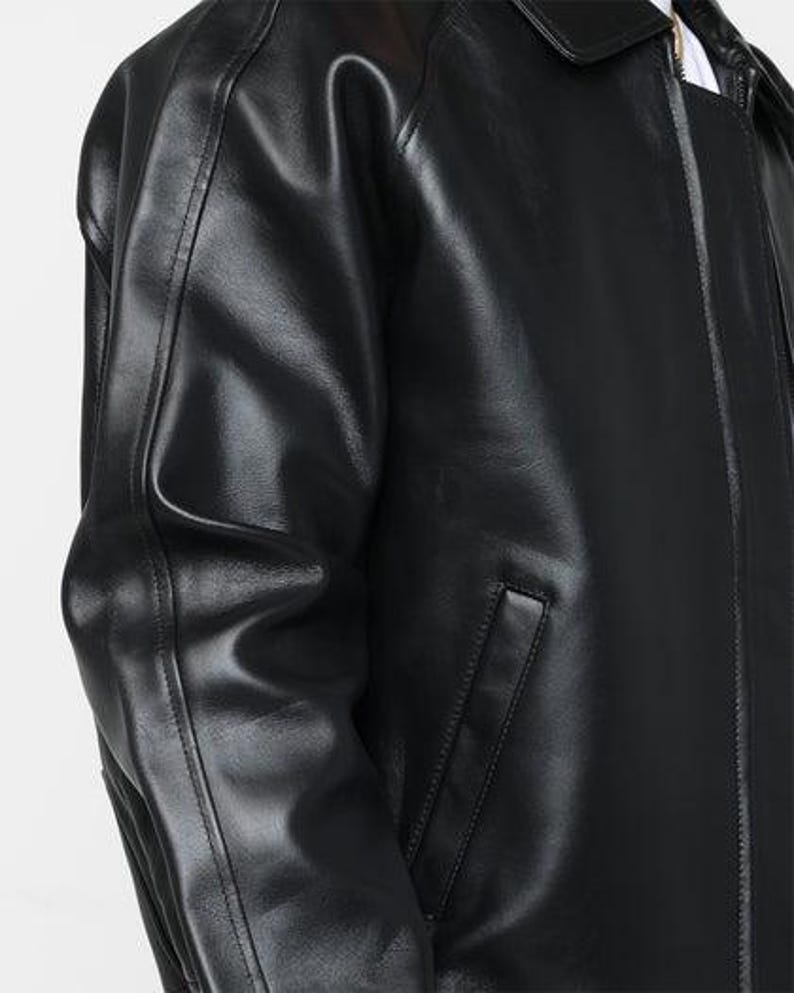 May include: A close-up of a black leather jacket. The jacket has a collar, a zipper, and a pocket on the front. The leather appears shiny and smooth.