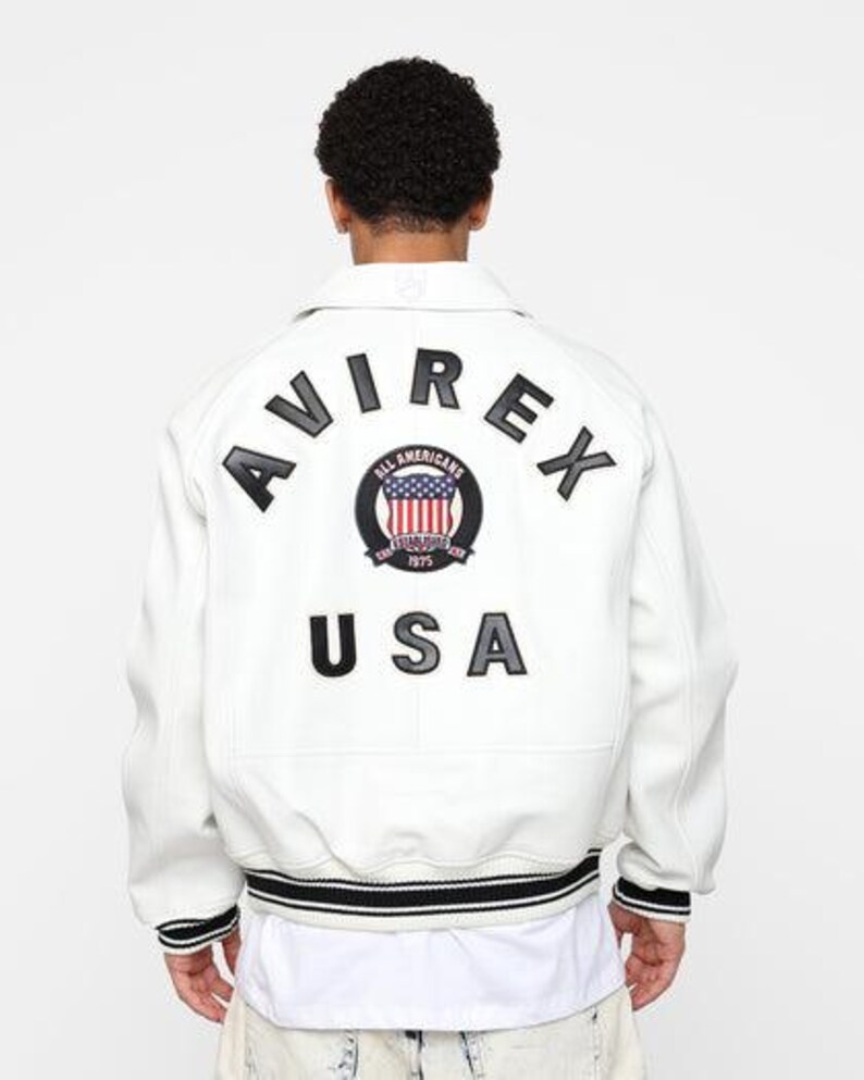 May include: White jacket with black and white striped trim and the text "AVIREX USA" on the back. The jacket features a circular patch with a US flag design and the words "ALL AMERICANS 1975".
