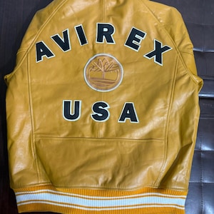 May include: Mustard yellow leather jacket with the text "AVIREX USA" in black and white. Features a circular patch with a tree design. The jacket has a ribbed knit hem with white and orange stripes.